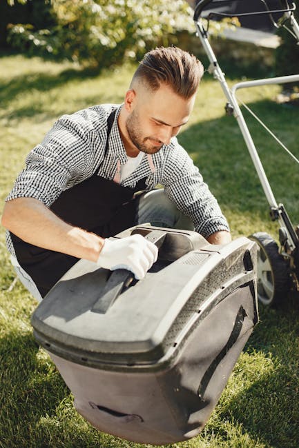 A man with light skin, styled brown hair, and a beard, wearing a checkered shirt and black apron, is kneeling on a well-maintained green lawn in a garden during daylight, inspecting a large black gras